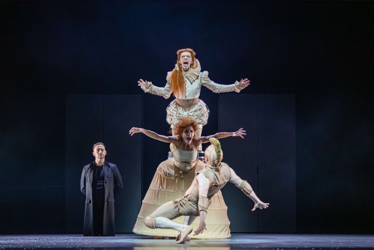 Four performers on stage; two are stacked atop each other in period costumes reminiscent of Mary, Queen of Scots, one is mid-air in front, and one stands in the background wearing a dark coat.