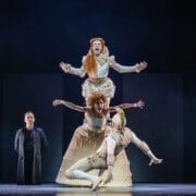 Four performers on stage; two are stacked atop each other in period costumes reminiscent of Mary, Queen of Scots, one is mid-air in front, and one stands in the background wearing a dark coat.