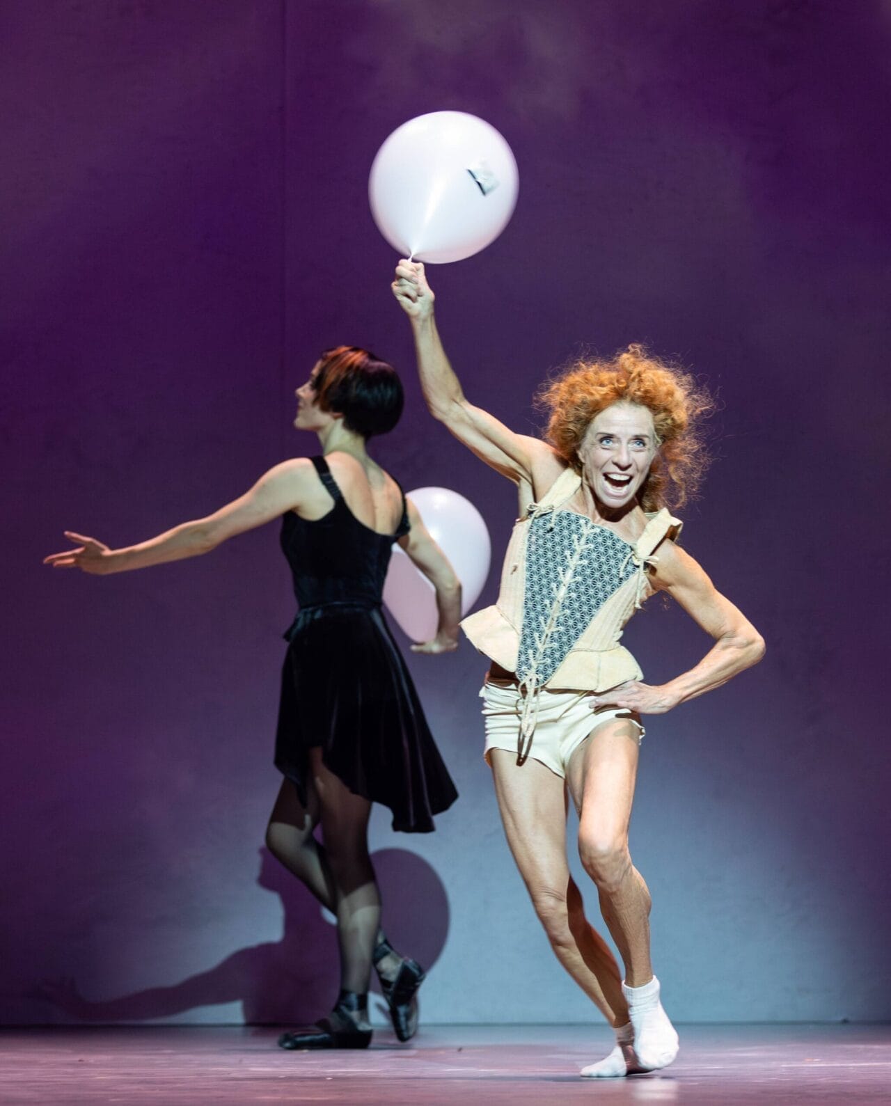 A performer in a corset and shorts holds a white balloon and poses energetically, while another evokes Mary, Queen of Scots, turning away in a dramatic black dress on stage.