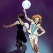 A performer in a corset and shorts holds a white balloon and poses energetically, while another evokes Mary, Queen of Scots, turning away in a dramatic black dress on stage.