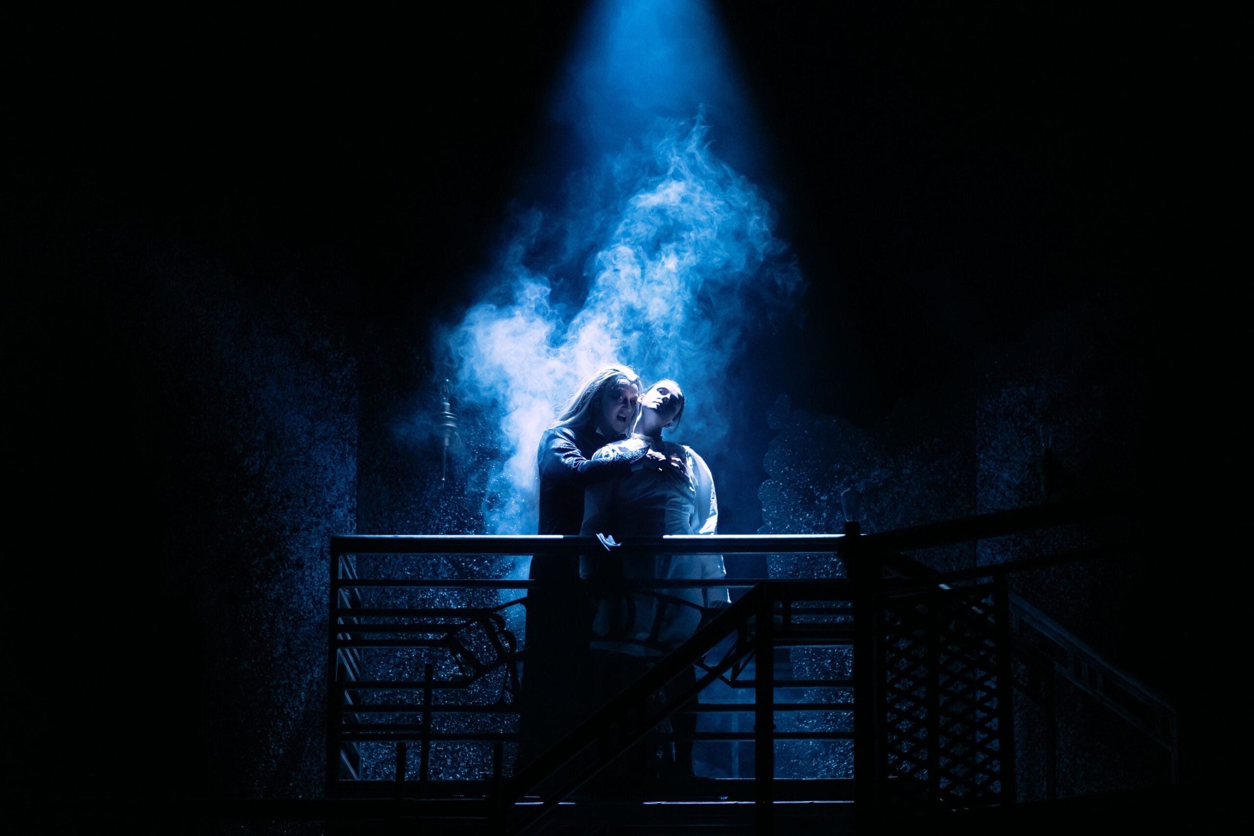 Two people stand on a staircase under a beam of blue stage light with dramatic smoke swirling around them in a dark setting, creating an atmosphere that gives you goosebumps.