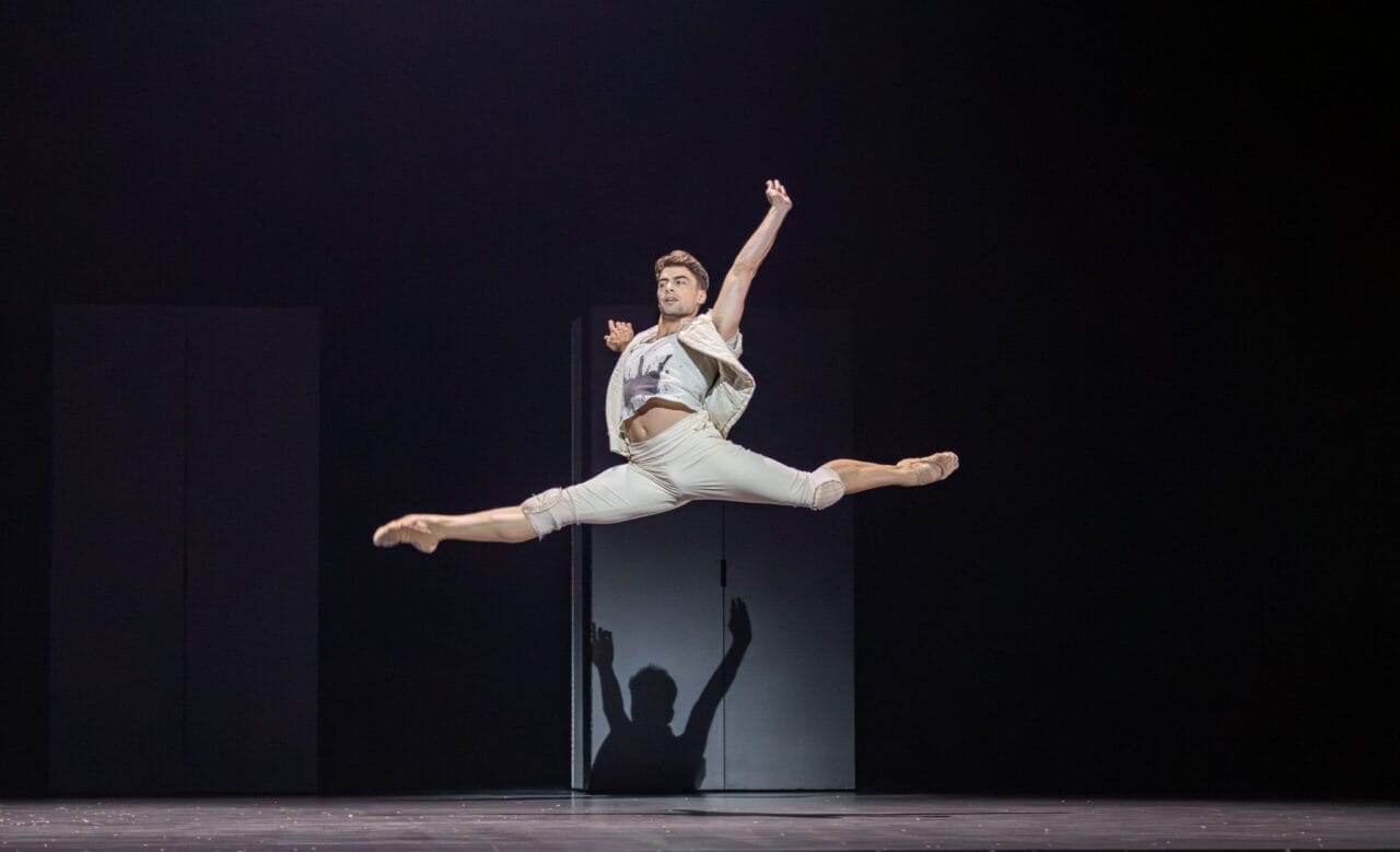 A male dancer performs a grand jeté on stage, leaping with arms and legs extended, evoking the dramatic spirit of Mary Queen of Scots in Scottish history, wearing a light-colored costume against a dark background.