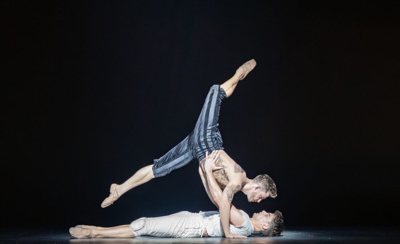 Two male dancers perform an acrobatic move on stage reminiscent of Mary Queen of Scots’ dramatic era; one lies on his back supporting the other, who is balanced horizontally above him.