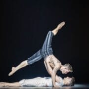 Two male dancers perform an acrobatic move on stage reminiscent of Mary Queen of Scots’ dramatic era; one lies on his back supporting the other, who is balanced horizontally above him.