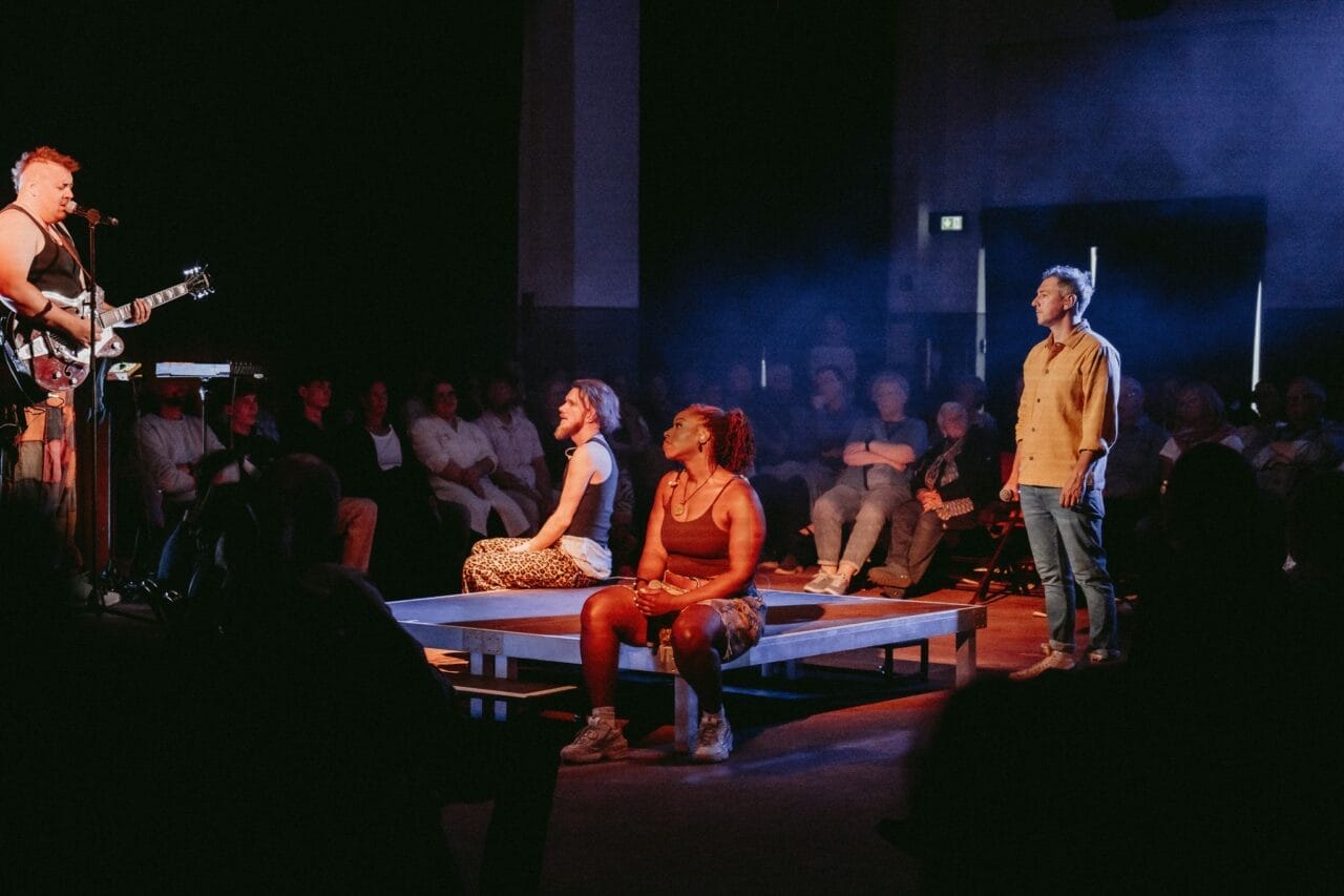 A musician plays guitar on stage while three people, two seated and one standing, face him—evoking a Half Man Half Bull dynamic. An audience watches in the background under dim lighting.