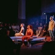 A musician plays guitar on stage while three people, two seated and one standing, face him—evoking a Half Man Half Bull dynamic. An audience watches in the background under dim lighting.