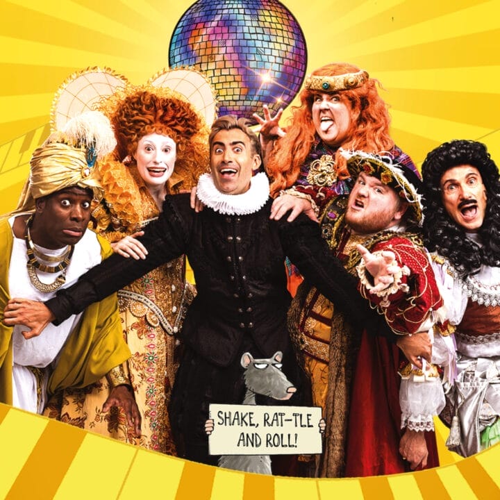 Seven people in colorful, elaborate historical costumes pose cheerfully in front of a disco ball on a yellow background with the text “Shake, Rat-tle and Roll!” below, celebrating the energy of a Horrible Histories live event.