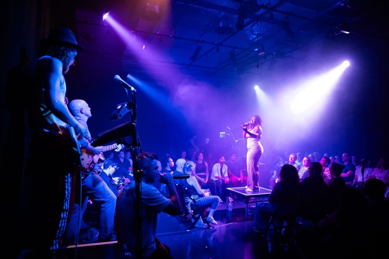 A woman sings on stage under purple lights with musicians playing beside her, while an audience—some captivated by a striking Minotaur masked performer—watches in a dimly lit theater.