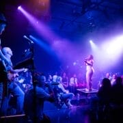 A woman sings on stage under purple lights with musicians playing beside her, while an audience—some captivated by a striking Minotaur masked performer—watches in a dimly lit theater.