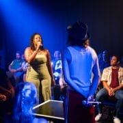 A woman sings into a microphone on stage under blue lighting, while another person in a hat stands opposite her—like a scene from Greek mythology, the audience surrounds them in a dimly lit room.