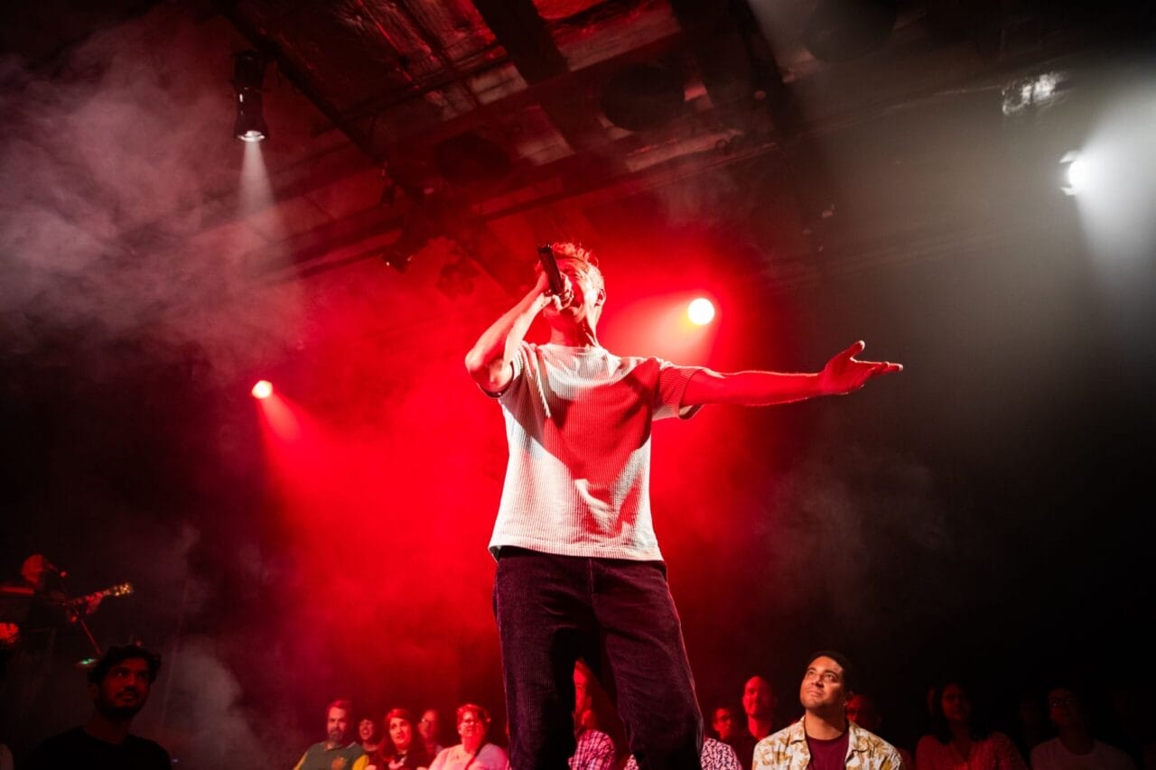 A person performs on stage with a microphone under red lights, channeling the energy of Half Man Half Bull, surrounded by an excited audience.