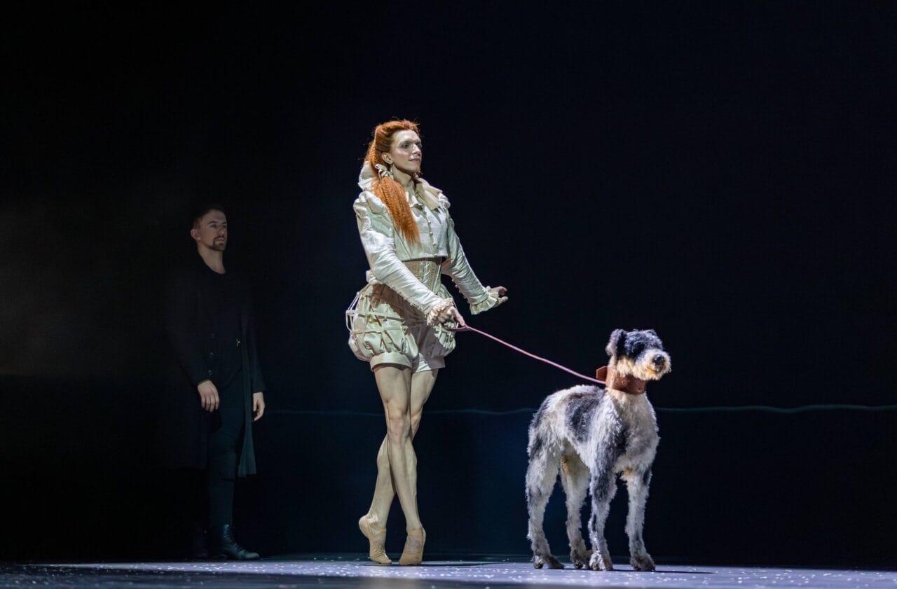 A performer in a light-colored costume and pointe shoes stands on stage holding a large dog on a leash, evoking the drama of Mary Queen of Scots, with another person in dark clothing in the background.