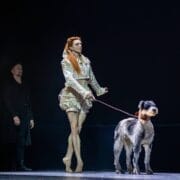 A performer in a light-colored costume and pointe shoes stands on stage holding a large dog on a leash, evoking the drama of Mary Queen of Scots, with another person in dark clothing in the background.
