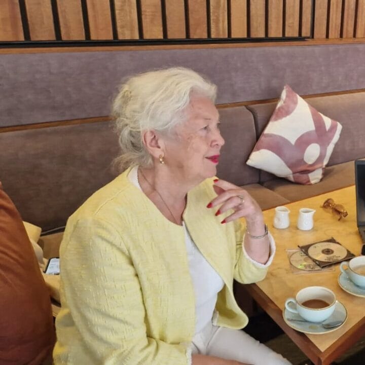 An elderly woman with white hair and a yellow jacket sits at a table with teacups and a CD, resting her hand on her chin in a cafe setting, enjoying the relaxed atmosphere of the Saturday Social—a wonderful example of Creative Ageing.