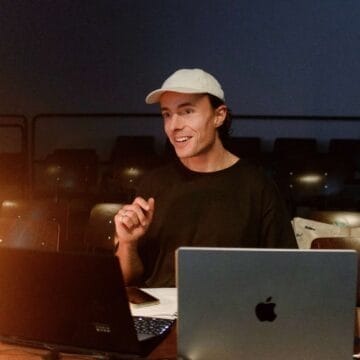 Edward Saunders, wearing a white cap and black shirt, sits at a table with two open laptops, gesturing while speaking in a dimly lit room with empty chairs in the background.