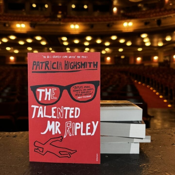 A copy of "The Talented Mr. Ripley" by Patricia Highsmith stands upright on a stage with a stack of books beside it; the empty theater auditorium hints at an awaiting Theatre Book Club gathering.