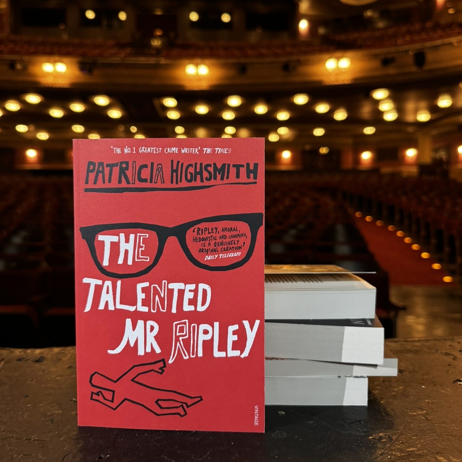 A copy of "The Talented Mr. Ripley" by Patricia Highsmith stands upright on a stage with a stack of books beside it; the empty theater auditorium hints at an awaiting Theatre Book Club gathering.