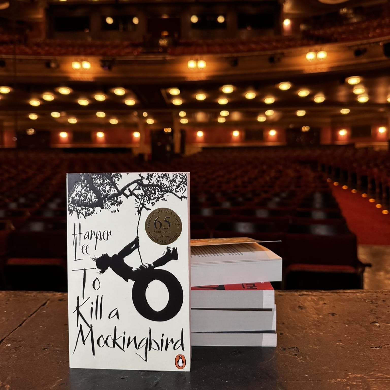 A copy of "To Kill a Mockingbird" by Harper Lee stands on a stage before stacked books, ready for discussion by the Theatre Book Club, with empty theater seats awaiting an audience in the background.