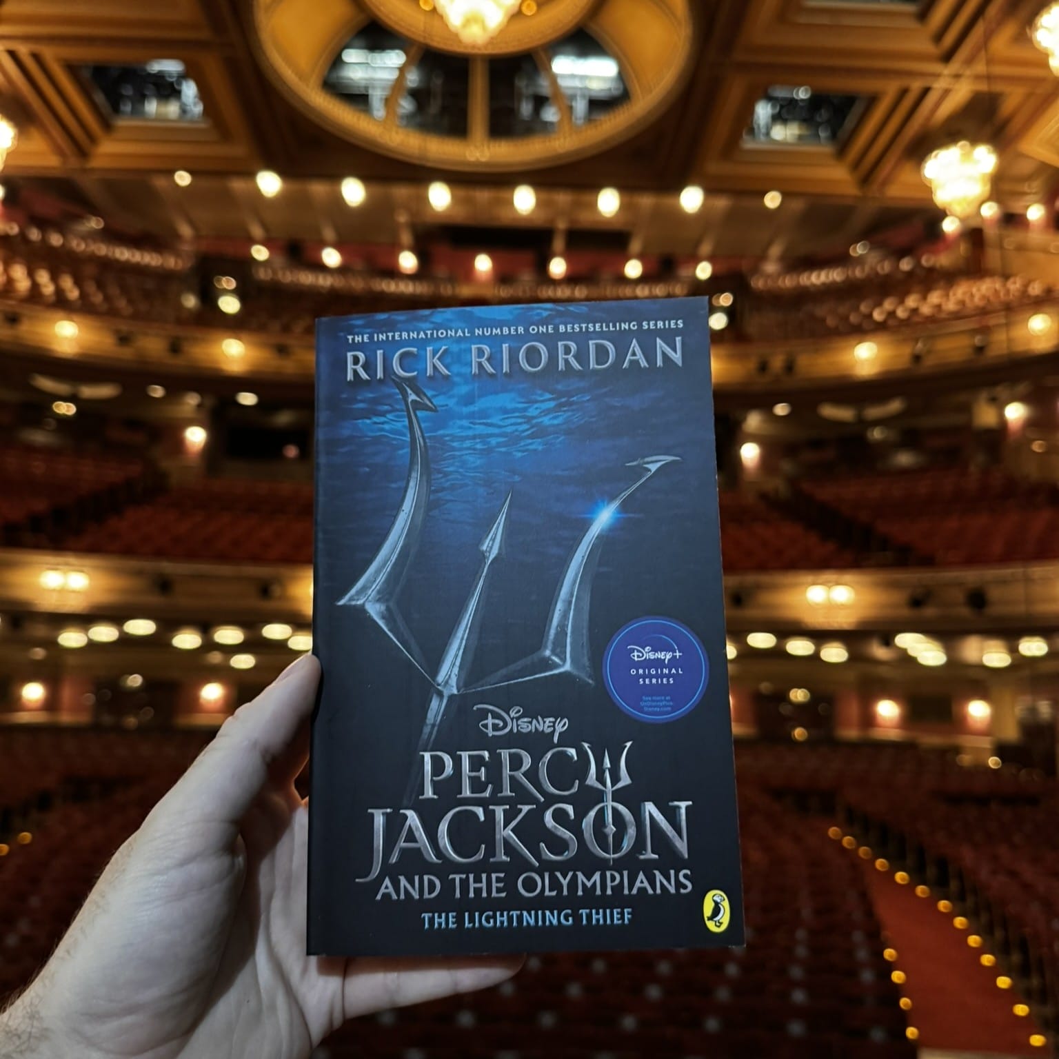 A hand holds a copy of "Percy Jackson and the Olympians: The Lightning Thief" by Rick Riordan in the foreground of an empty, ornate theater—perfect for a Theatre Book Club gathering.