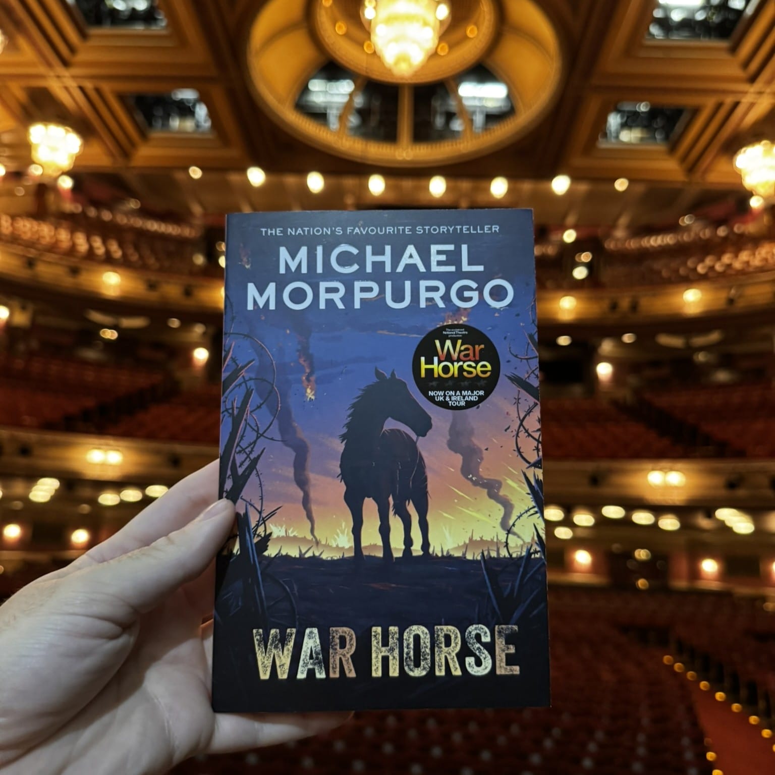 A hand holds a copy of "War Horse" by Michael Morpurgo, perfect for any Theatre Book Club, with a theater auditorium and chandeliers softly blurred in the background.