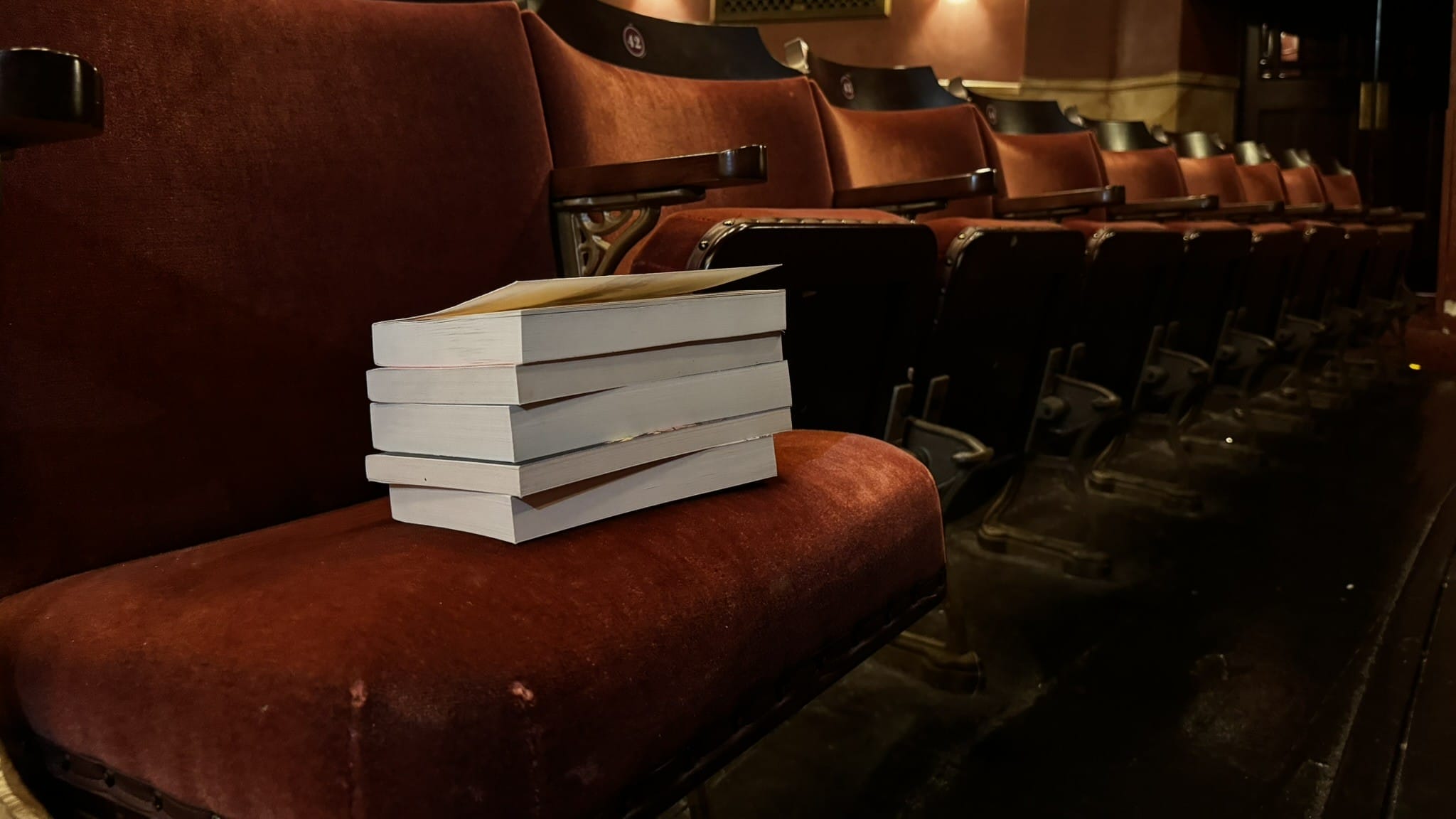 A stack of books sits on a red velvet theater seat in an empty auditorium, inviting a Book Club gathering amidst ornate walls and dim lighting.