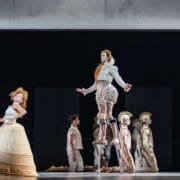 Performers in stylized, historical costumes stand and kneel on stage; one person, portraying Mary Queen of Scots, is elevated on stilts, surrounded by others in matching attire against a dark backdrop.