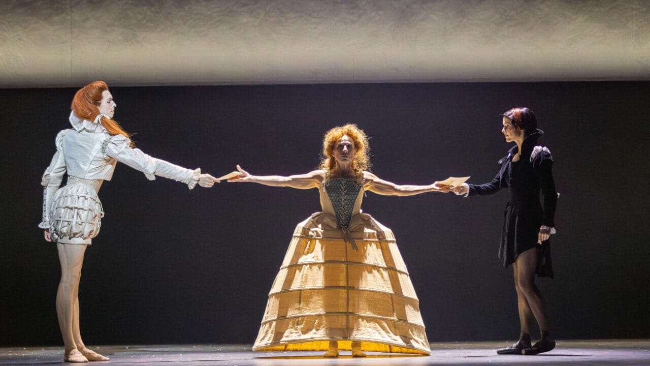 Three performers in period costumes stand on stage; the center figure, representing Mary Queen of Scots, with outstretched arms connects the two others, each holding one of her hands.