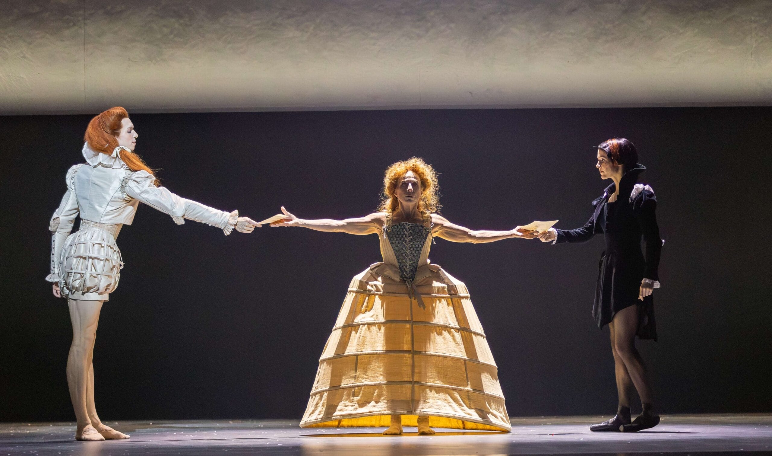 Three performers in period costumes stand on stage; the center figure, representing Mary Queen of Scots, with outstretched arms connects the two others, each holding one of her hands.