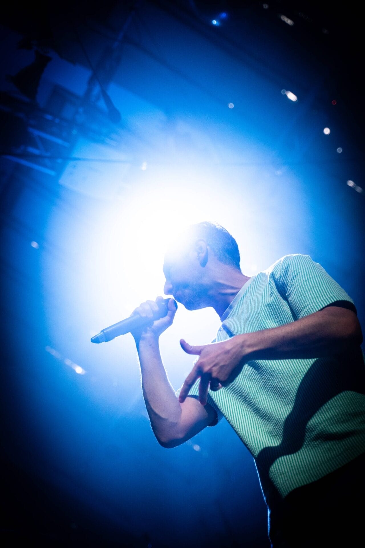 A person holding a microphone performs on stage under bright blue and white lighting, embodying the spirit of Half Man Half Bull.