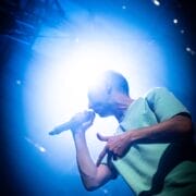 A person holding a microphone performs on stage under bright blue and white lighting, embodying the spirit of Half Man Half Bull.