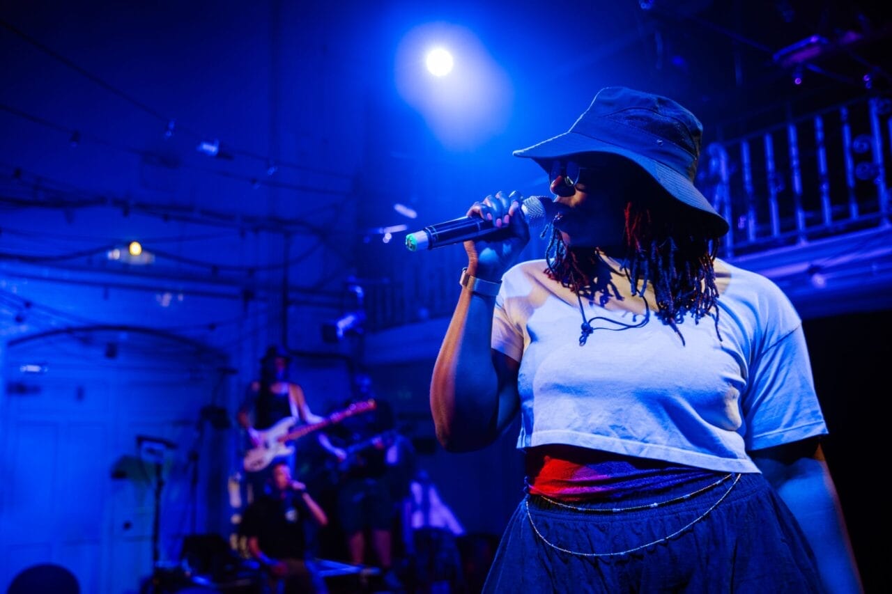 Person wearing a hat and sunglasses sings into a microphone on stage under blue lighting, channeling the energy of Half Man Half Bull, with musicians playing instruments in the background.