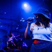 Person wearing a hat and sunglasses sings into a microphone on stage under blue lighting, channeling the energy of Half Man Half Bull, with musicians playing instruments in the background.