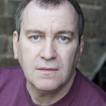 Mark Hadfield, a middle-aged man with short thinning hair and blue eyes, wears a maroon shirt and looks directly at the camera with a neutral expression. A brick wall is visible in the background.