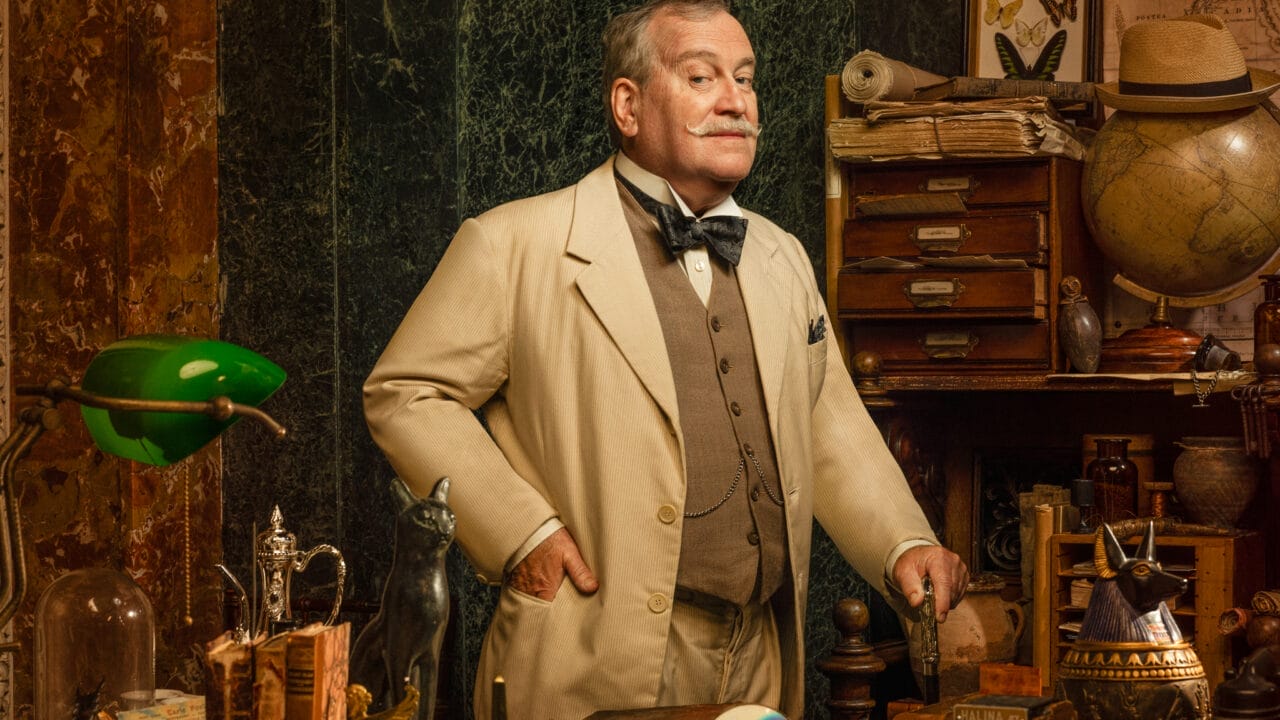 Mark Hadfield, dressed in a beige suit, stands in a cluttered study filled with books, scientific instruments, and curiosities, with shelves and drawers lining the background.