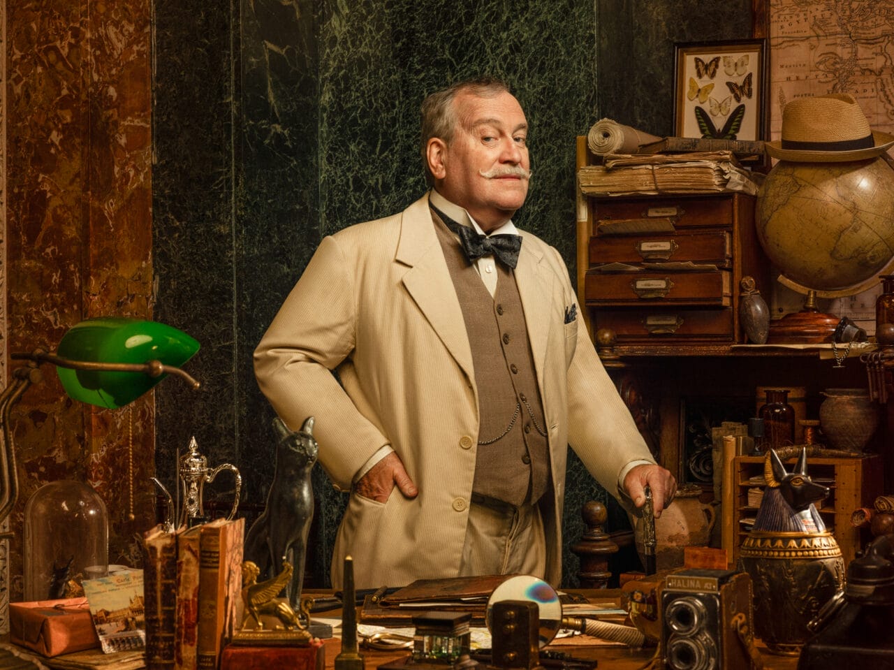 Mark Hadfield, dressed in a beige suit, stands in a cluttered study filled with books, scientific instruments, and curiosities, with shelves and drawers lining the background.