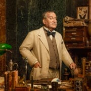 Mark Hadfield, dressed in a beige suit, stands in a cluttered study filled with books, scientific instruments, and curiosities, with shelves and drawers lining the background.