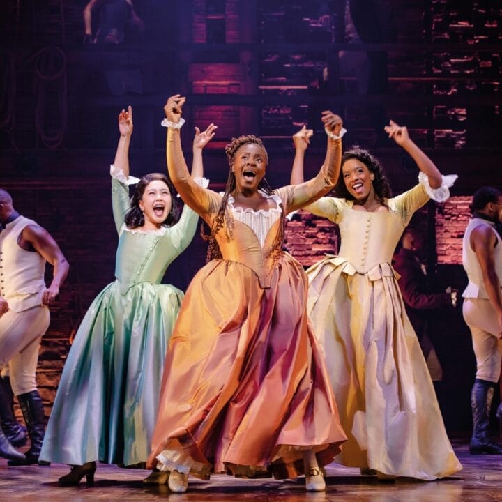 Three women in period costumes sing and gesture energetically on stage, with other 25 and under performers in colonial-era attire in the background, during a lively theatrical performance.