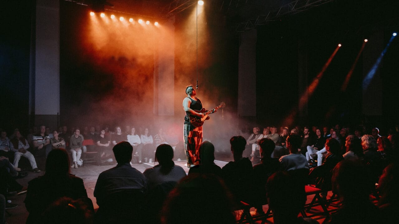 A musician, reminiscent of Half Man Half Bull, performs on a circular stage surrounded by an audience, with colorful stage lighting and smoke effects.