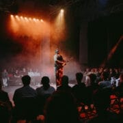 A musician, reminiscent of Half Man Half Bull, performs on a circular stage surrounded by an audience, with colorful stage lighting and smoke effects.