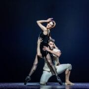 Two ballet dancers perform on stage; the woman in a black outfit stands en pointe with one arm raised, evoking Mary Queen of Scots, while the man in light attire kneels and supports her.