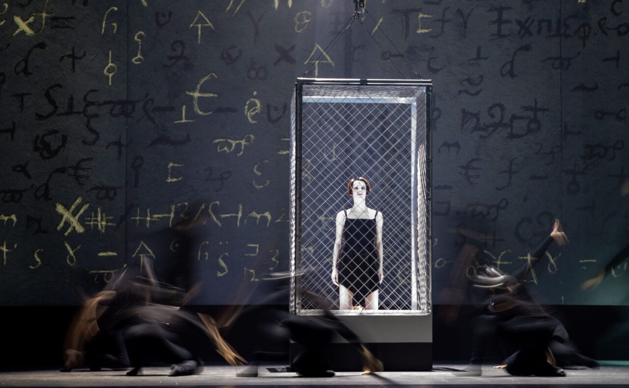 A person in a black outfit and white face makeup stands inside a wire cage on stage, evoking Mary Queen of Scots, with dark blurred figures moving around and abstract symbols on the background wall.
