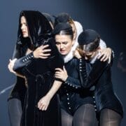 A group of performers in black velvet costumes huddle together on stage, evoking the intrigue of Mary Queen of Scots, with one wearing a hooded cloak and others in ruffs and hats, all under dramatic lighting.