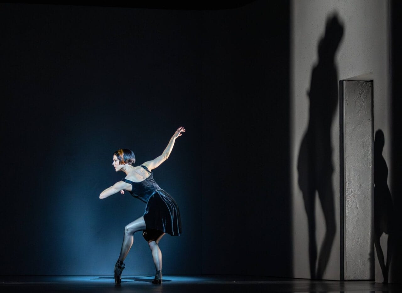 A dancer in a black dress poses dramatically on stage, her silhouette evoking the regal presence of Mary, Queen of Scots, with a large shadow cast on the white wall beside her.