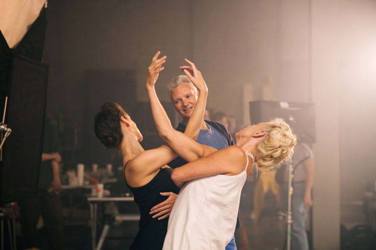 Two women practice a dance pose while a man stands nearby watching and smiling, as if observing Mary, Queen of Scots herself perfecting her moves, in a studio setting with equipment in the background.
