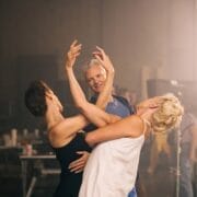 Two women practice a dance pose while a man stands nearby watching and smiling, as if observing Mary, Queen of Scots herself perfecting her moves, in a studio setting with equipment in the background.