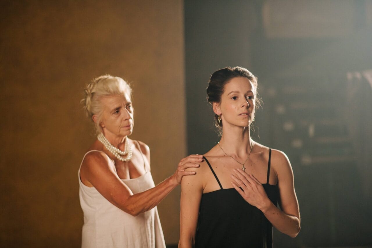 An older woman in a white dress rests her hand on the shoulder of a younger woman in black, who stands with her hand on her chest—both looking serious, evoking the solemn grace of Mary Queen of Scots.