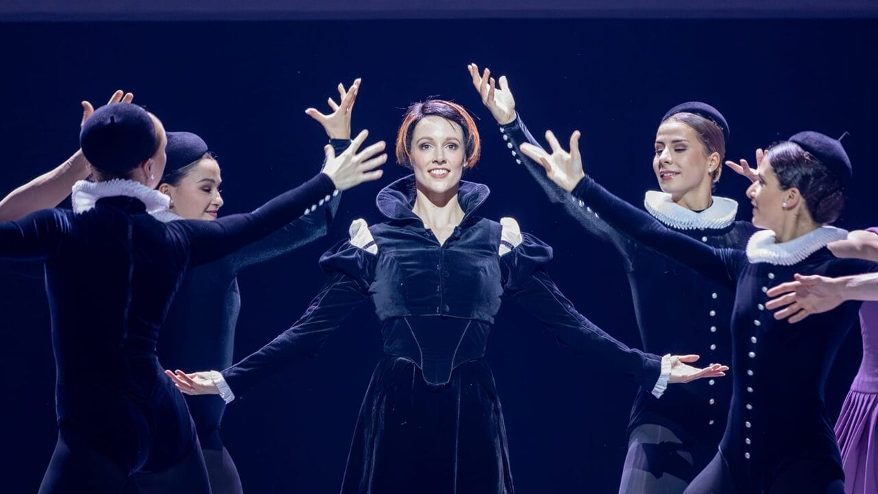 A group of ballet dancers performs on stage, with a central dancer in black costume evoking Mary Queen of Scots, surrounded by others in matching outfits, their arms extended in expressive poses that echo Scottish history.