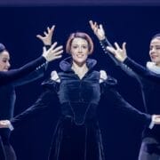 A group of ballet dancers performs on stage, with a central dancer in black costume evoking Mary Queen of Scots, surrounded by others in matching outfits, their arms extended in expressive poses that echo Scottish history.