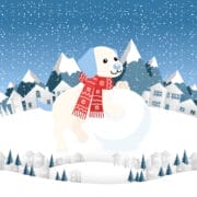 Cartoon dog wearing a red winter scarf stands on a large snowball in a snowy landscape with houses, trees, and mountains in the background. Snow is falling from the sky.