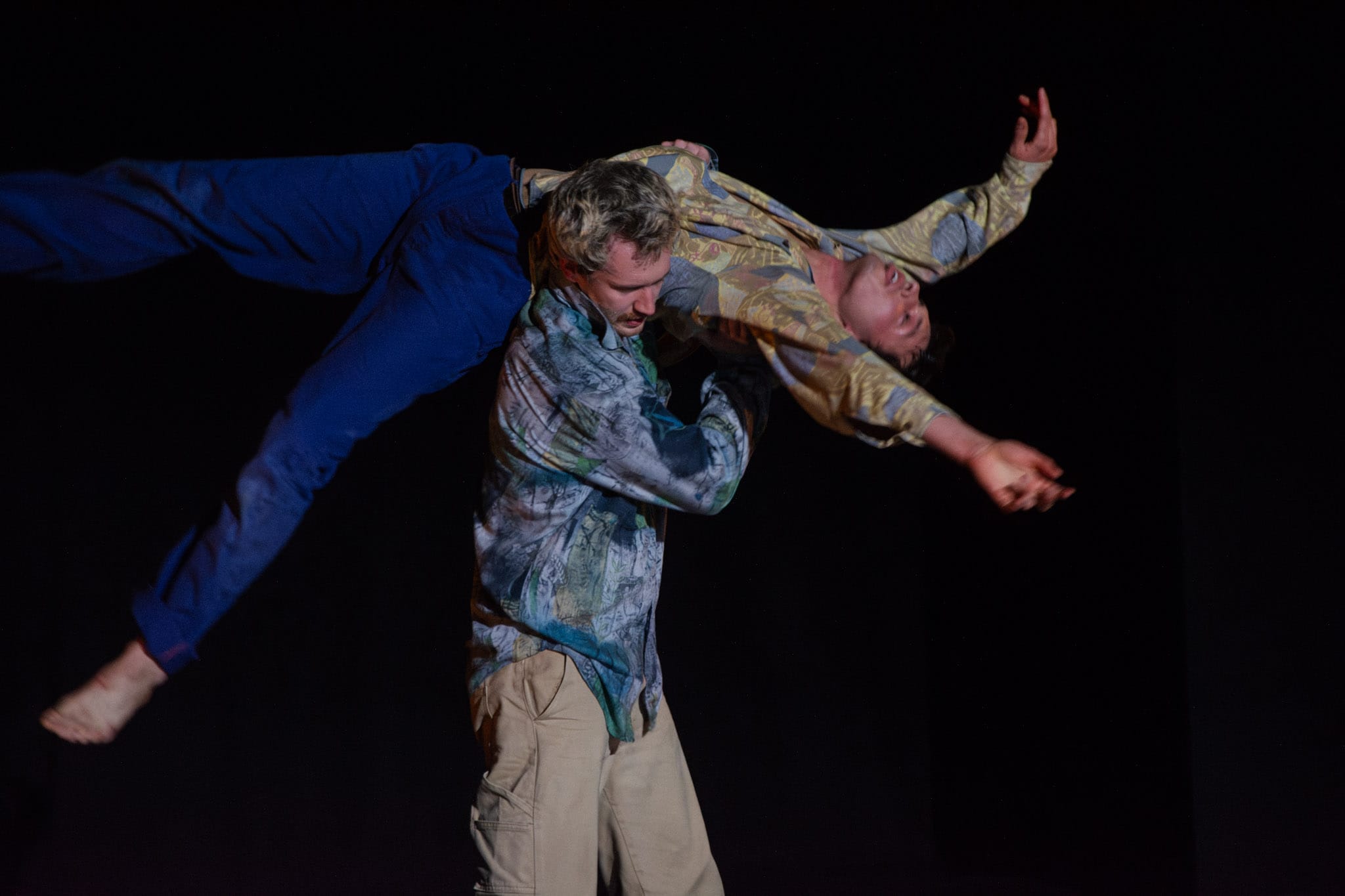 Two people perform an acrobatic move; one person lifts and balances the other horizontally over their shoulders against a dark background, showcasing striking dance innovations that push the boundaries of Dance Horizons.